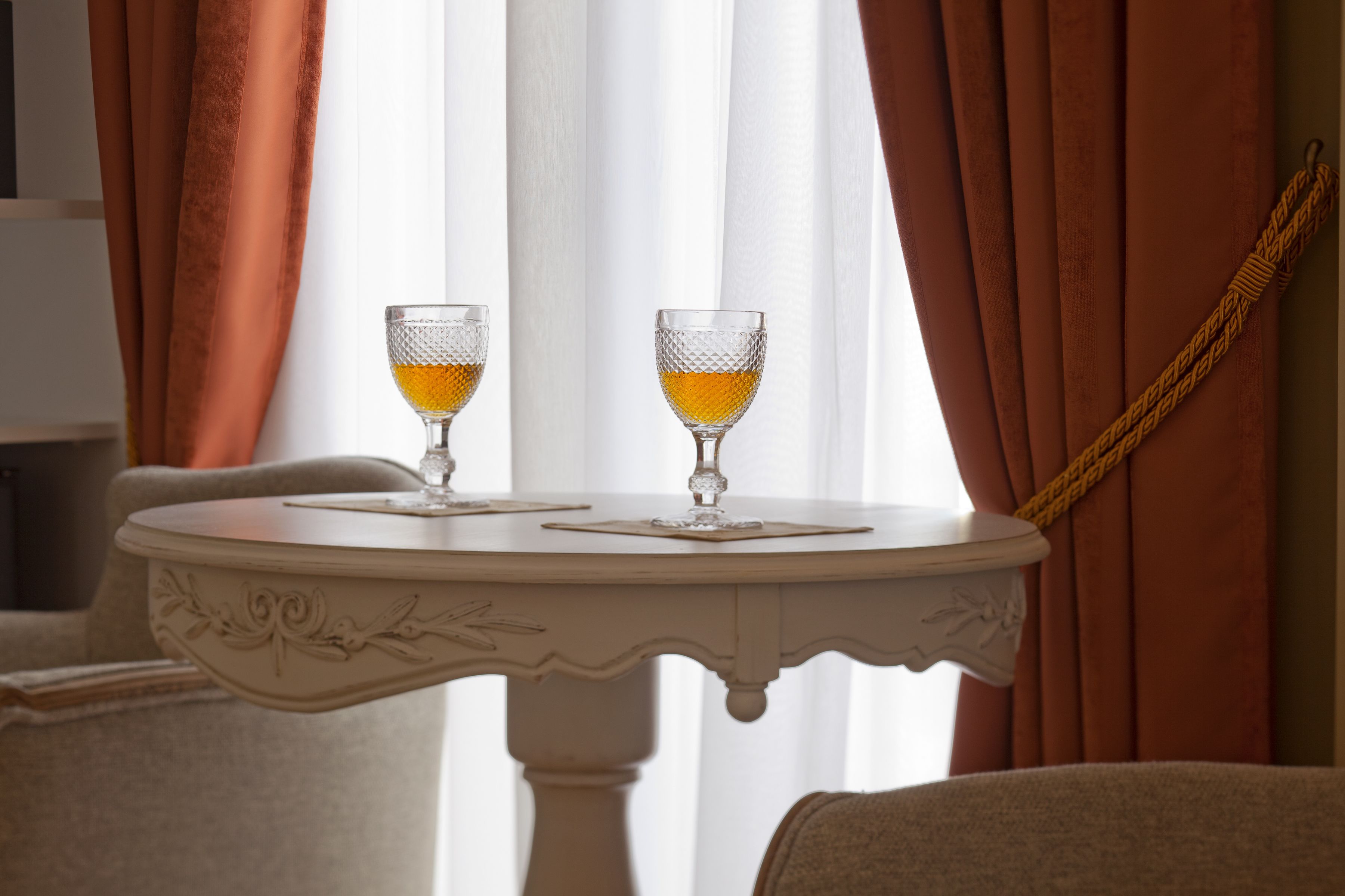 A round white table with two crystal glasses on a white tablecloth, set against a window with brown curtains.