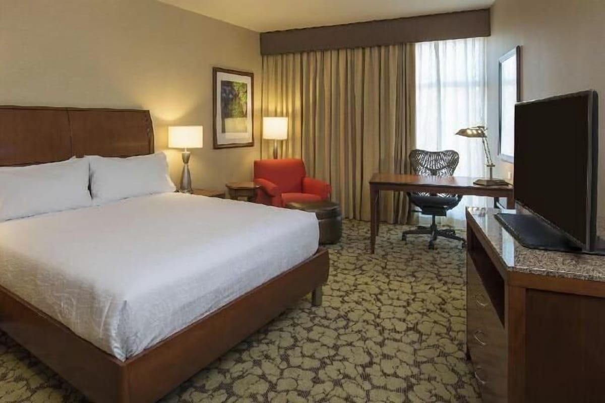 Une chambre d’hôtel avec un grand lit, deux tables de chevet avec des lampes, un bureau avec une chaise, une télévision et une fenêtre avec des rideaux.