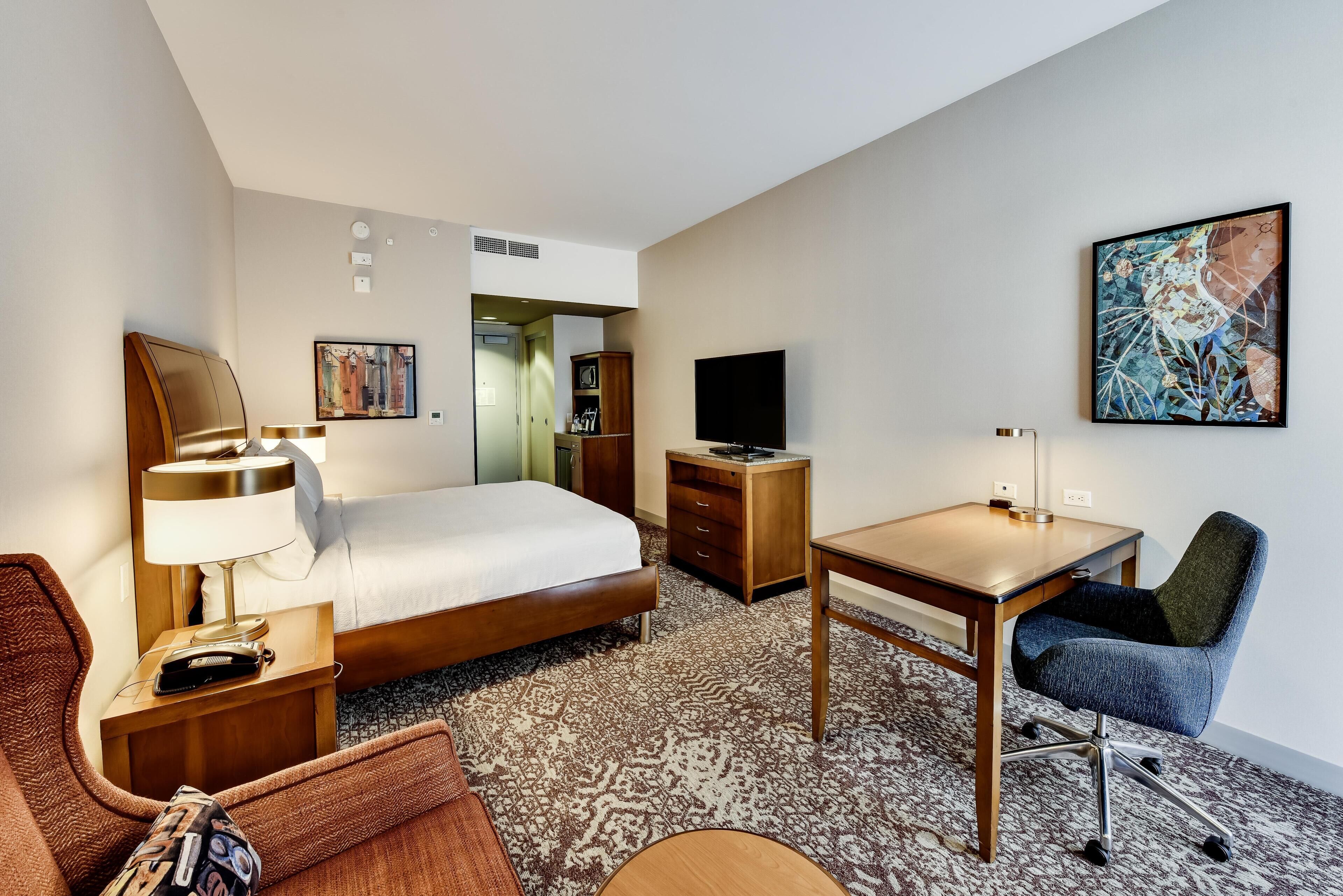 A hotel room with a bed, a desk, a chair, a television, and artwork on the walls.