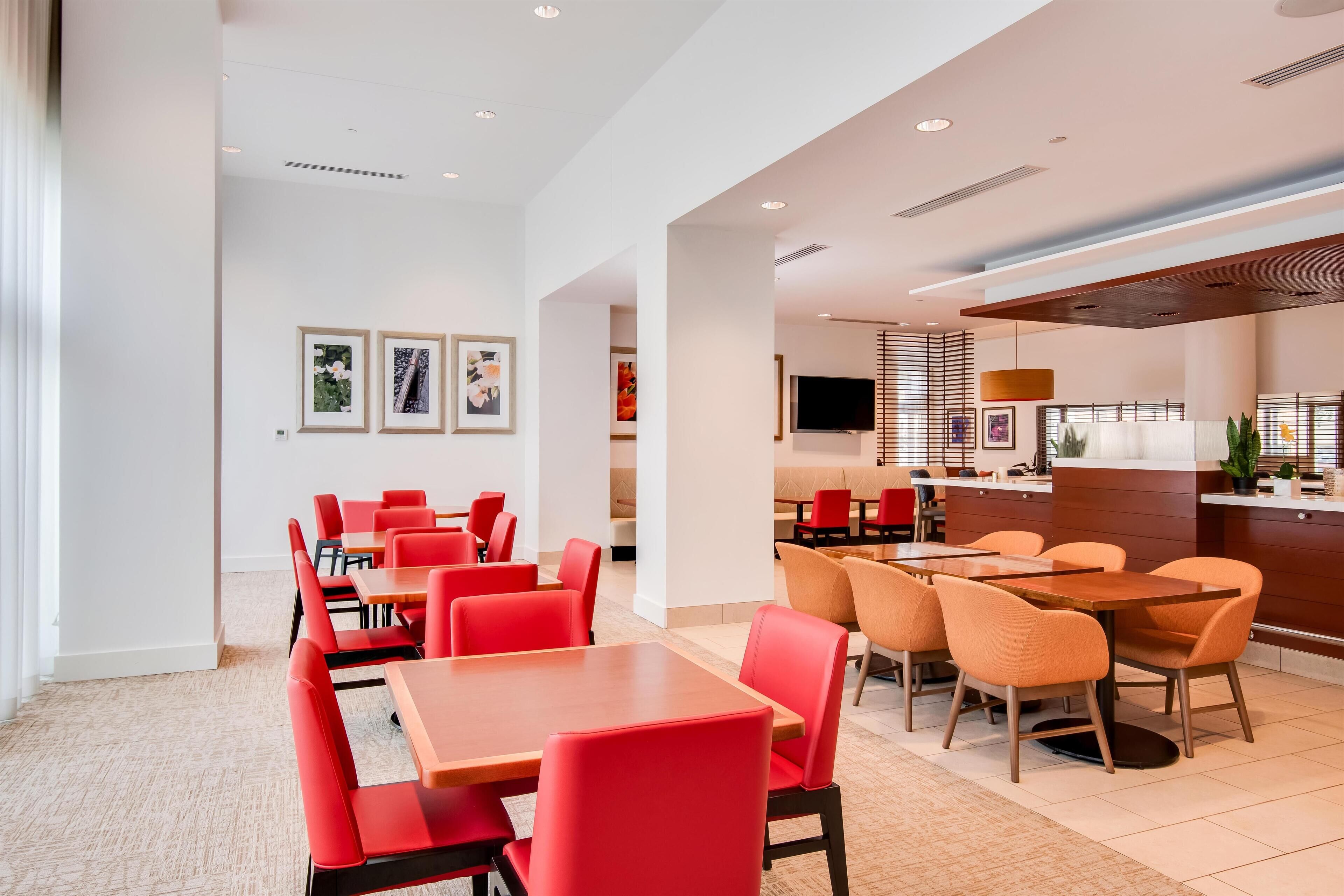 A modern dining area with red and orange chairs, a wooden table, and a kitchen area with a TV and counter.