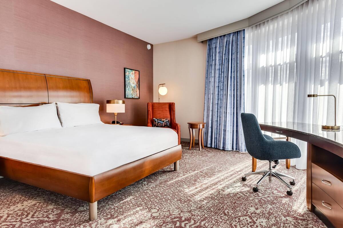 Ett hotellrum med en stor säng, ett skrivbord, en stol, ett litet bord och ett fönster med gardiner.