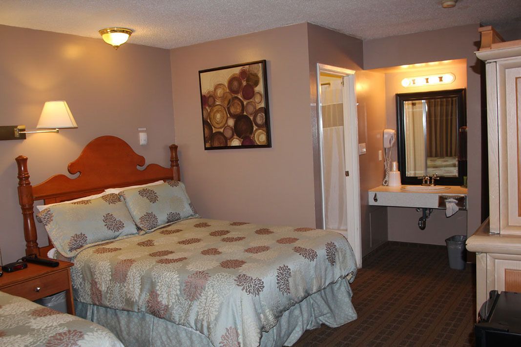 A hotel room with a wooden bed, a bedside lamp, a wall-mounted painting, a bathroom with a sink and mirror, and a trash bin.