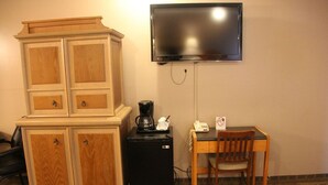 Una habitación con un armario de madera, un refrigerador negro pequeño, una cafetera, una mesa con una silla y un televisor montado.
