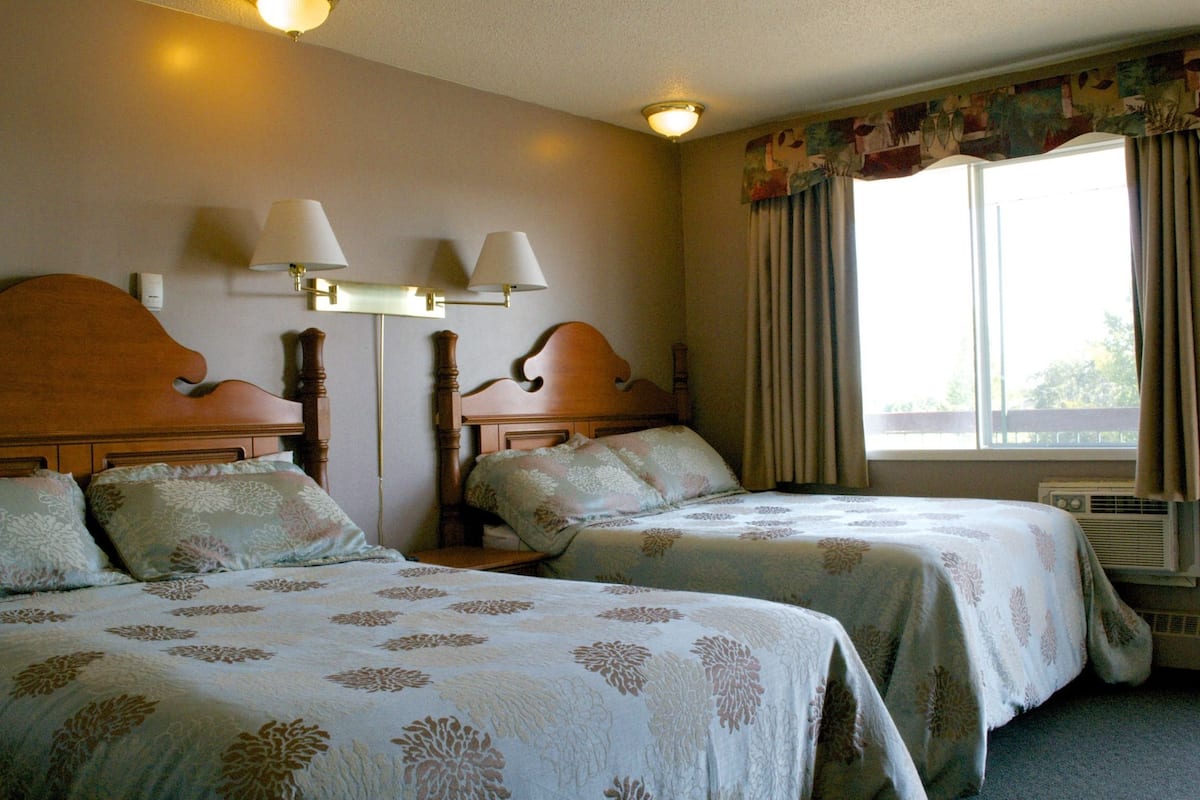 Ein Hotelzimmer mit zwei Betten, jeweils mit blumigem Bettbezug, einem Holz-Kopfteil, einem Nachttisch, einer Lampe und einem Fenster mit Vorhängen.