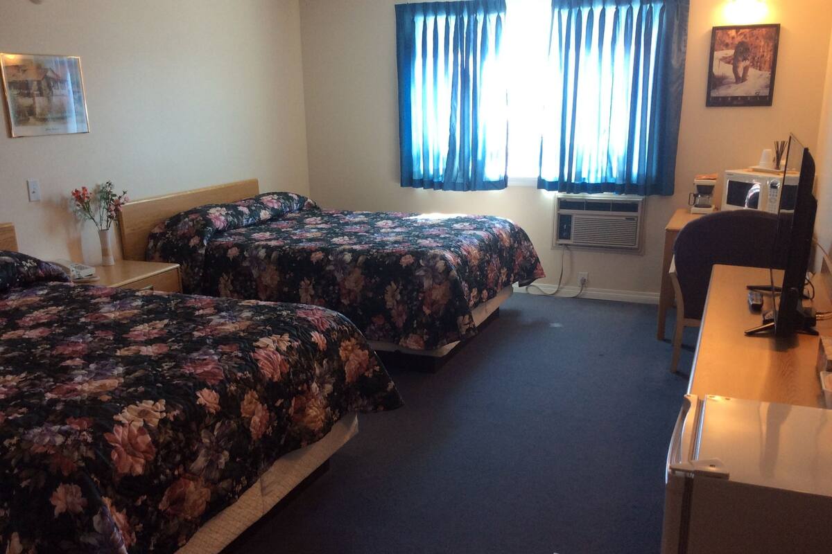 Habitación de hotel con dos camas, un escritorio, una silla, una mesita peque ña, un equipo de aire acondicionado y una ventana con cortinas.