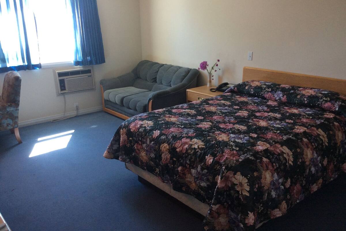 Un dormitorio con cama, sofá, mesita de noche, un jarrón con flores y ventana con cortinas.