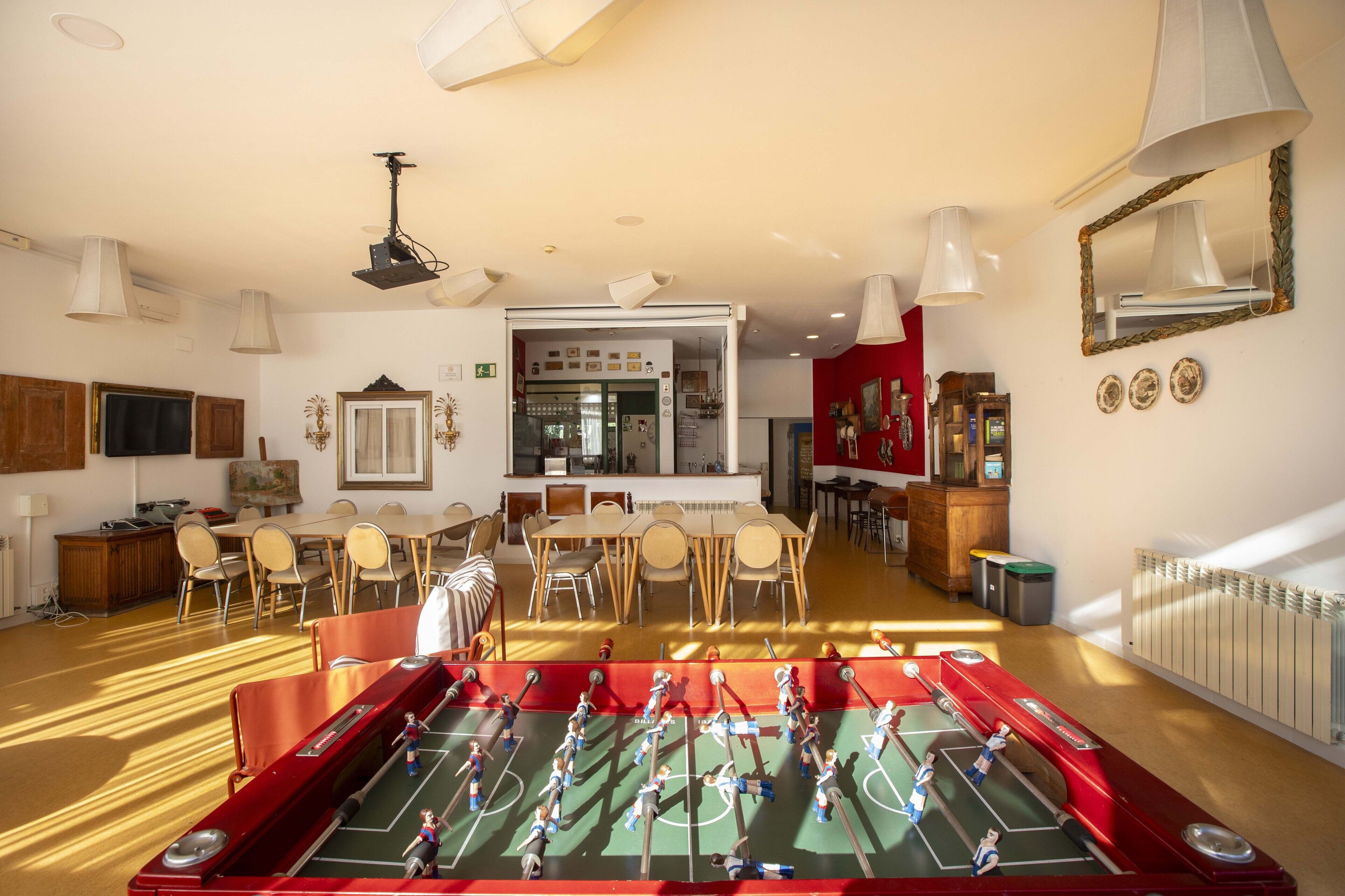 Una habitación amplia con mesa de fútbol, comedor con sillas y una mesa, y cocina visible a través de una puerta abierta.