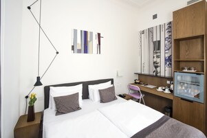 Habitación de hotel con cama, mesita de noche, lámpara, obra de arte, escritorio, silla y minibar.