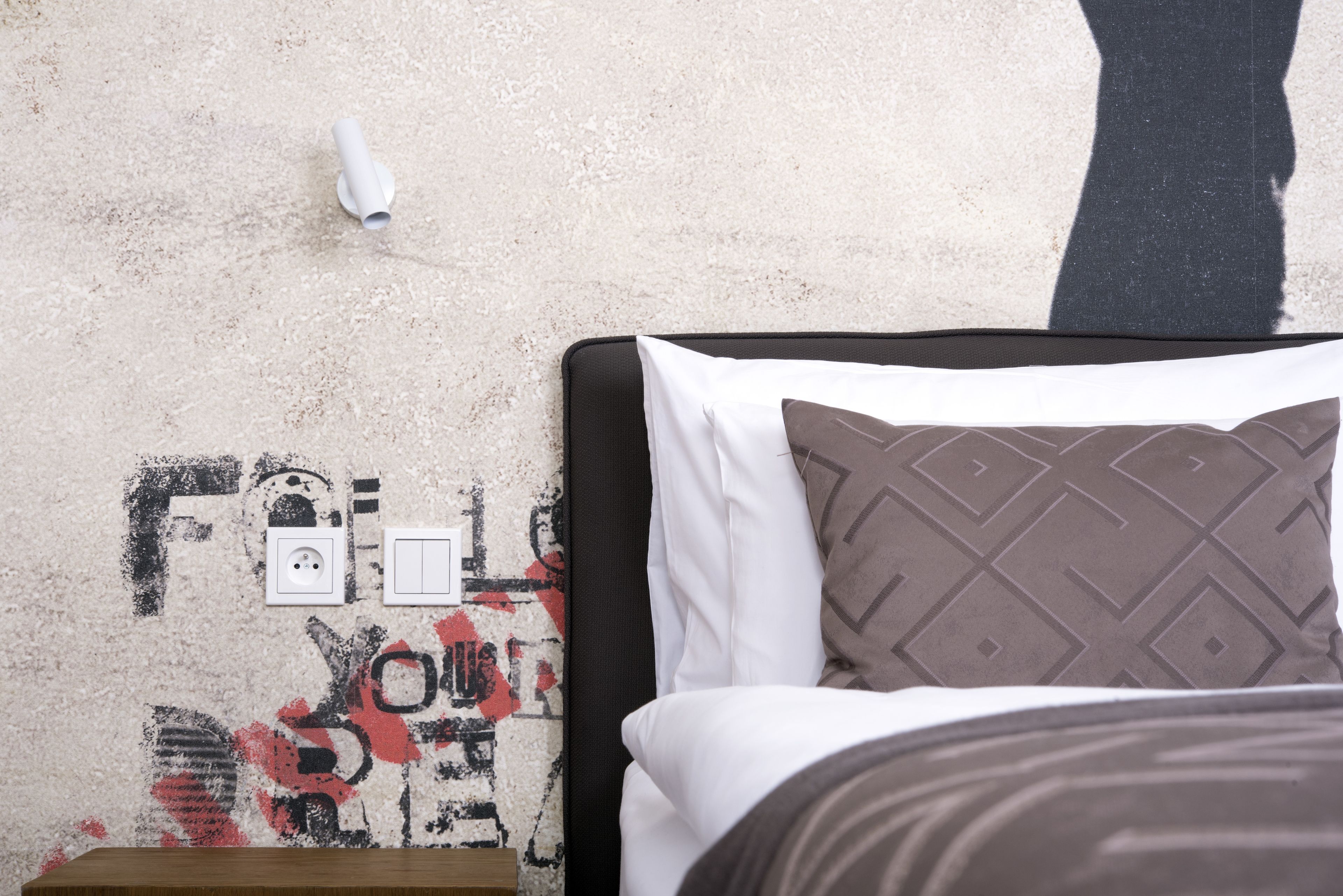 Un lit avec un oreiller à motifs, un mur blanc orné d’un dessin de style graffiti et une prise de courant.