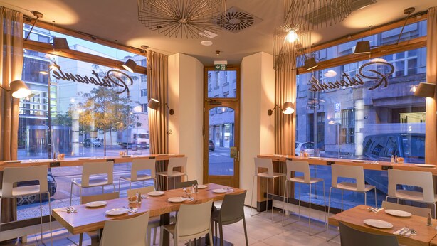 A restaurant with a modern interior, featuring wooden tables and chairs, large windows with curtains, and a view of the street outside. - Golden Crown (Prague)