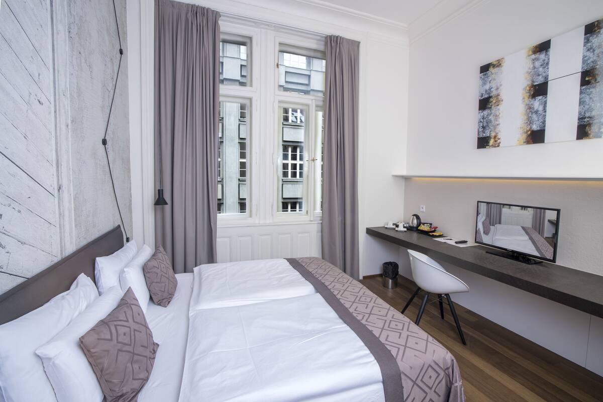 Habitación de hotel con cama, escritorio, silla, televisor y ventana con cortinas.