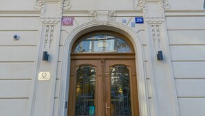 A building with a wooden double door, a plaque, and a window with a blue curtain. - Golden Crown (Prague)