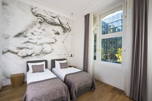 A bedroom with two beds, a large wall mural, a window with curtains, and a view of buildings outside.