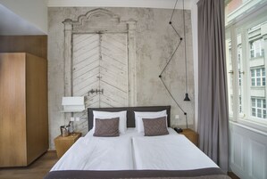 Uma cama de casal com lençóis brancos e duas almofadas estampadas, uma mesa de cabeceira de madeira com luminária e uma janela grande com cortinas.