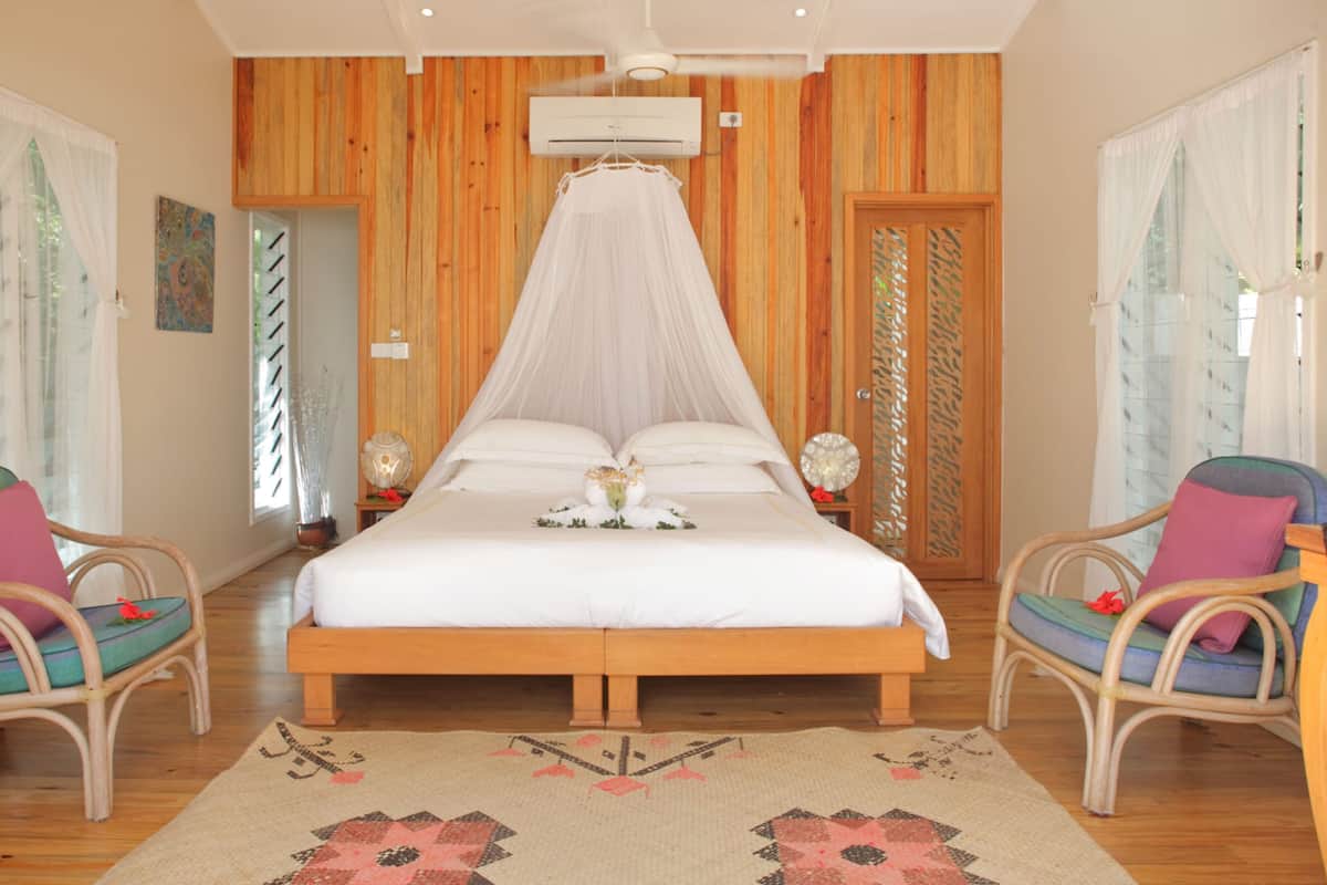 Ein Schlafzimmer mit einer holzverkleideten Wand, einem Himmelbett, zwei Korbstühlen, einem großen Teppich und einer Wand-Klimaanlage.