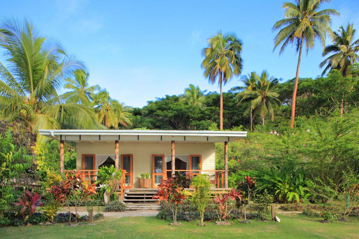 Ein Bungalow mit überdachter Veranda, umgeben von üppiger tropischer Vegetation und Palmen.