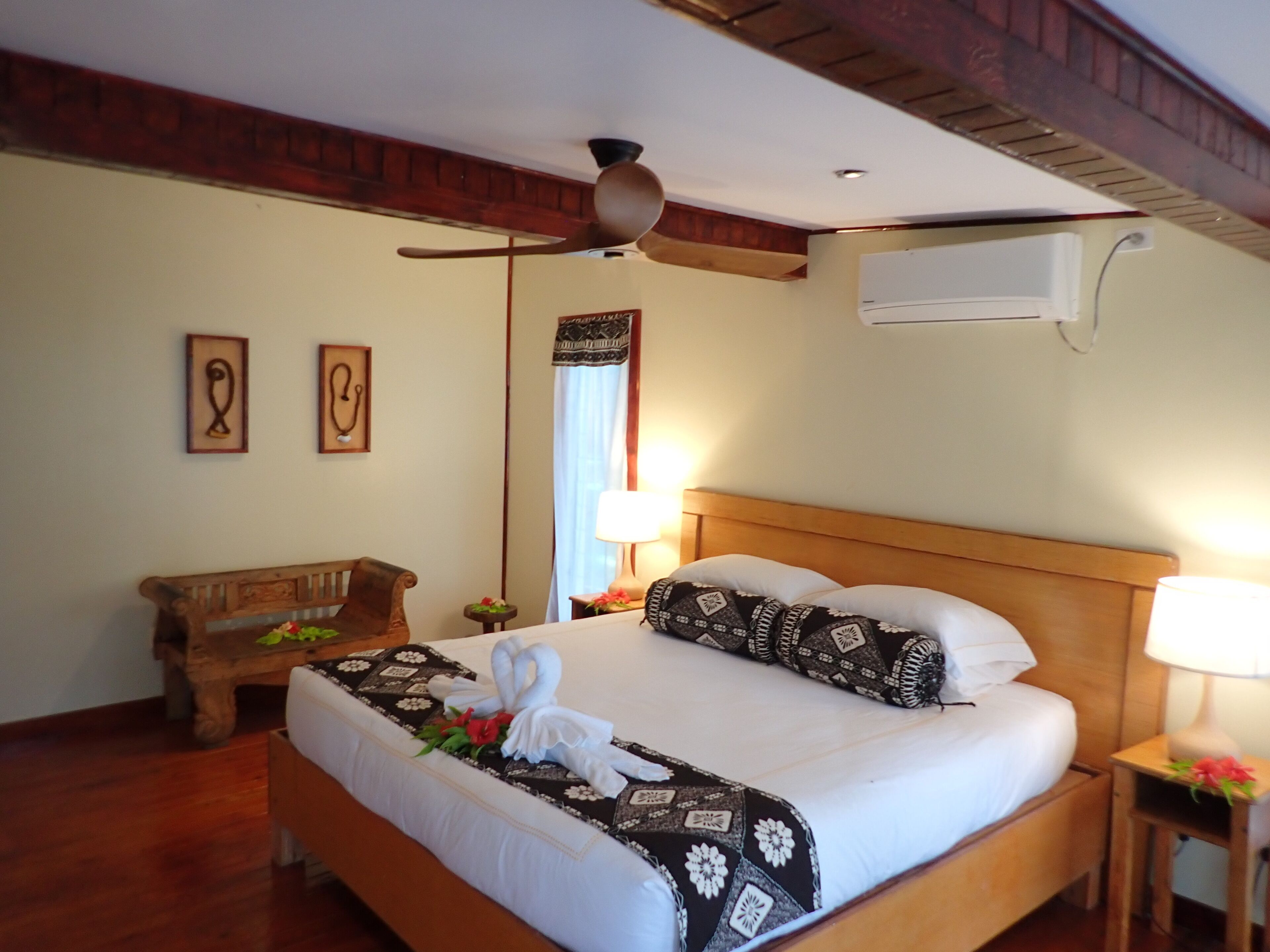 Una habitación de hotel con una cama y un banco de madera, un ventilador de techo y un equipo de aire acondicionado.