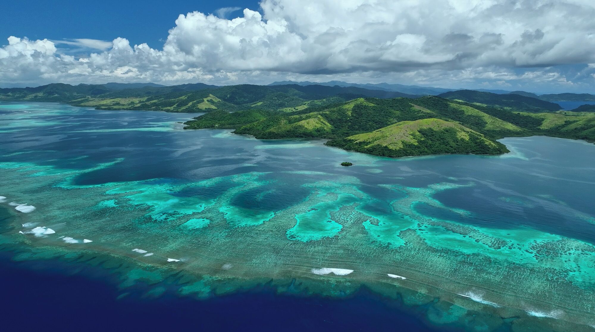 Vue aérienne d’une île tropicale aux eaux turquoise et limpides, bordée de collines verdoyantes.