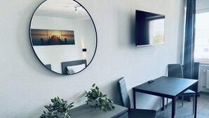 Una habitación de hotel moderna con un espejo redondo, un televisor de pantalla plana, una mesa de comedor con sillas y una planta en maceta.