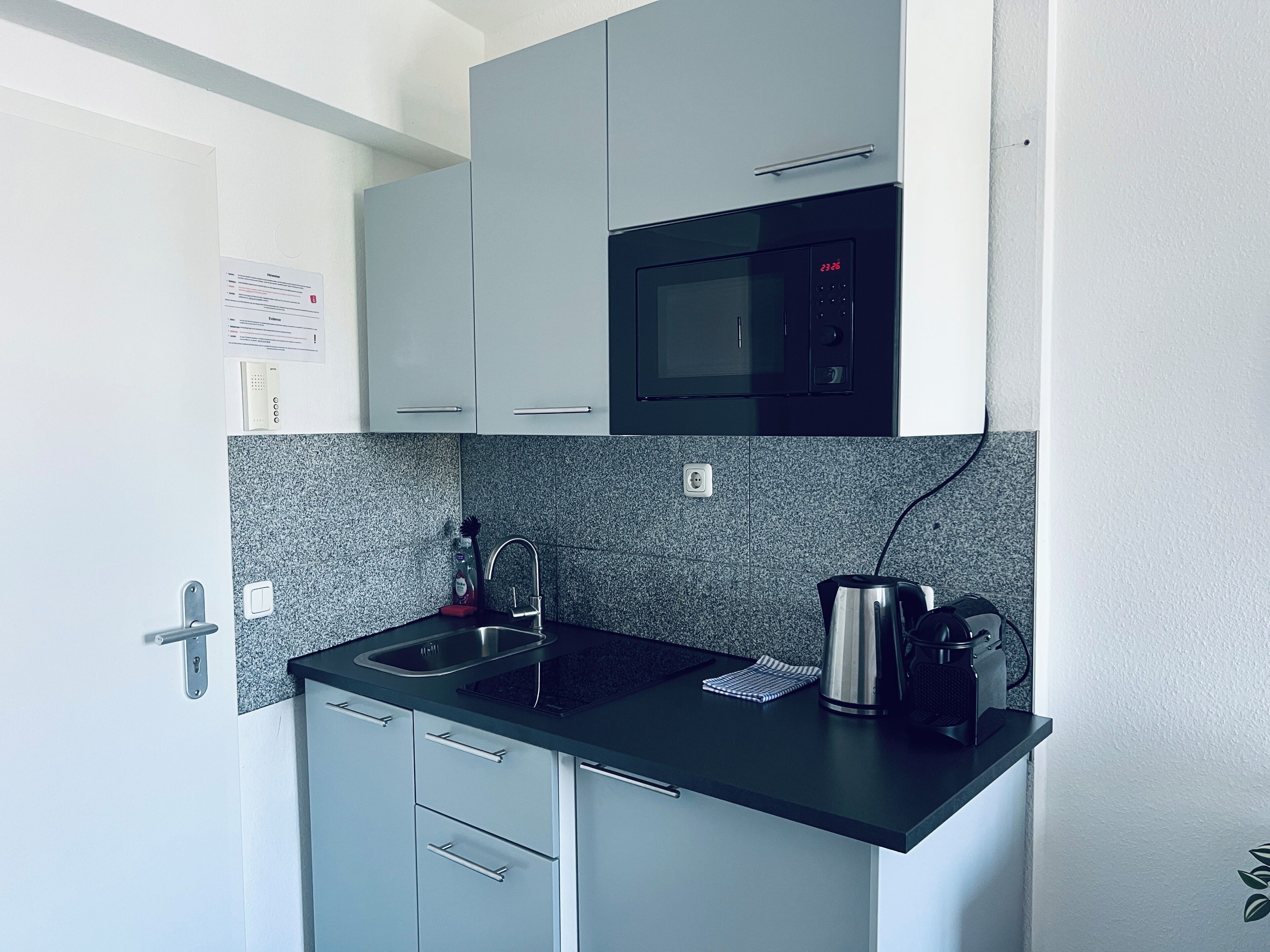 Een compacte keuken met grijze kastjes, een zwart aanrecht, een roestvrijstalen spoelbak, een magnetron en een waterkoker.