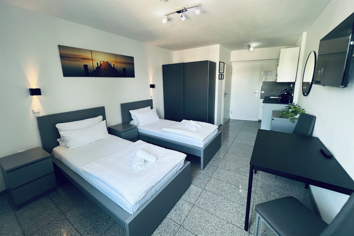 A modern hotel room with two beds, a desk, a kitchenette, and a bathroom.