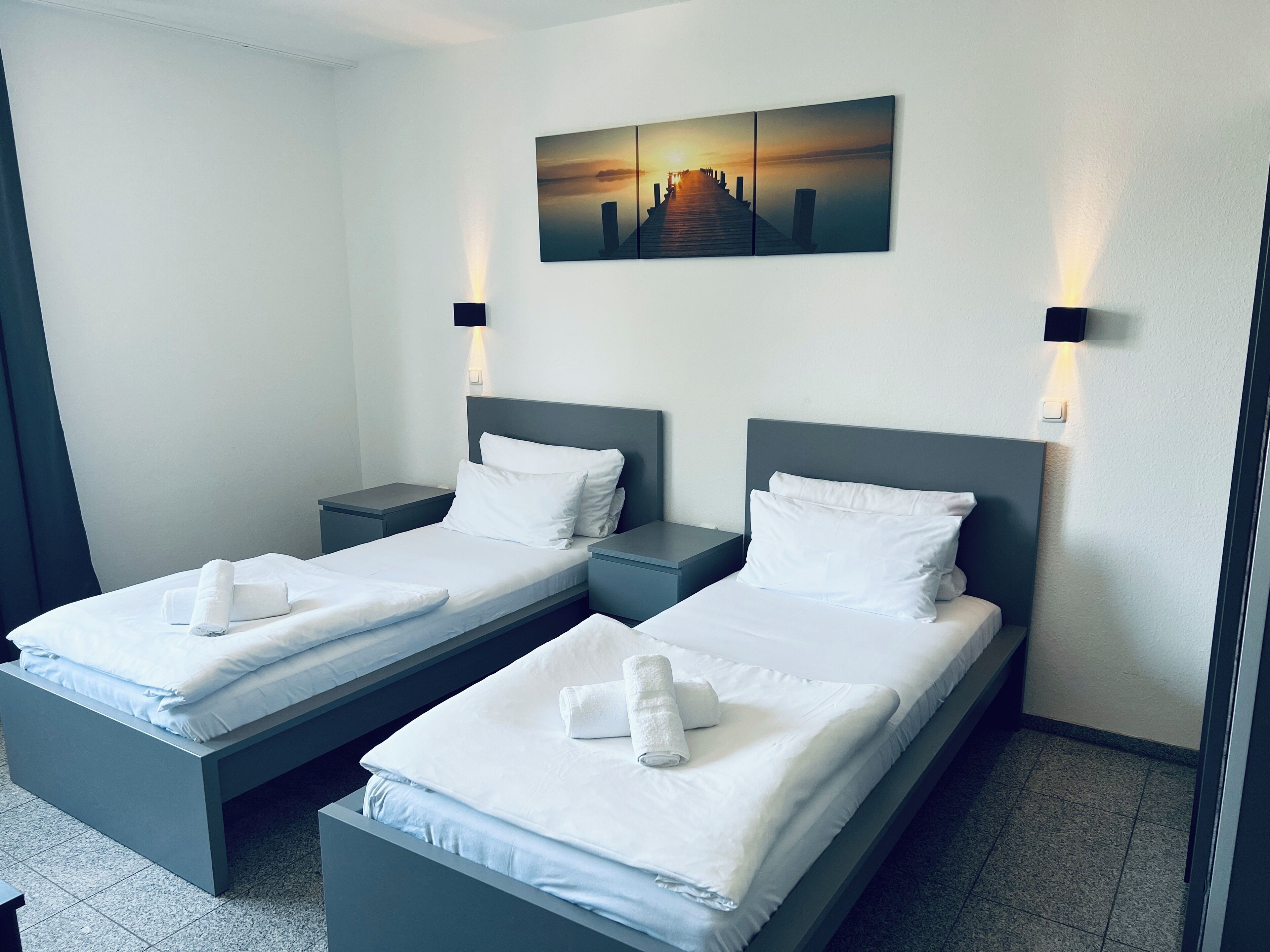 A hotel room with two beds, each with white linens and towels, a painting of a sunset over a pier, and wall-mounted lights.