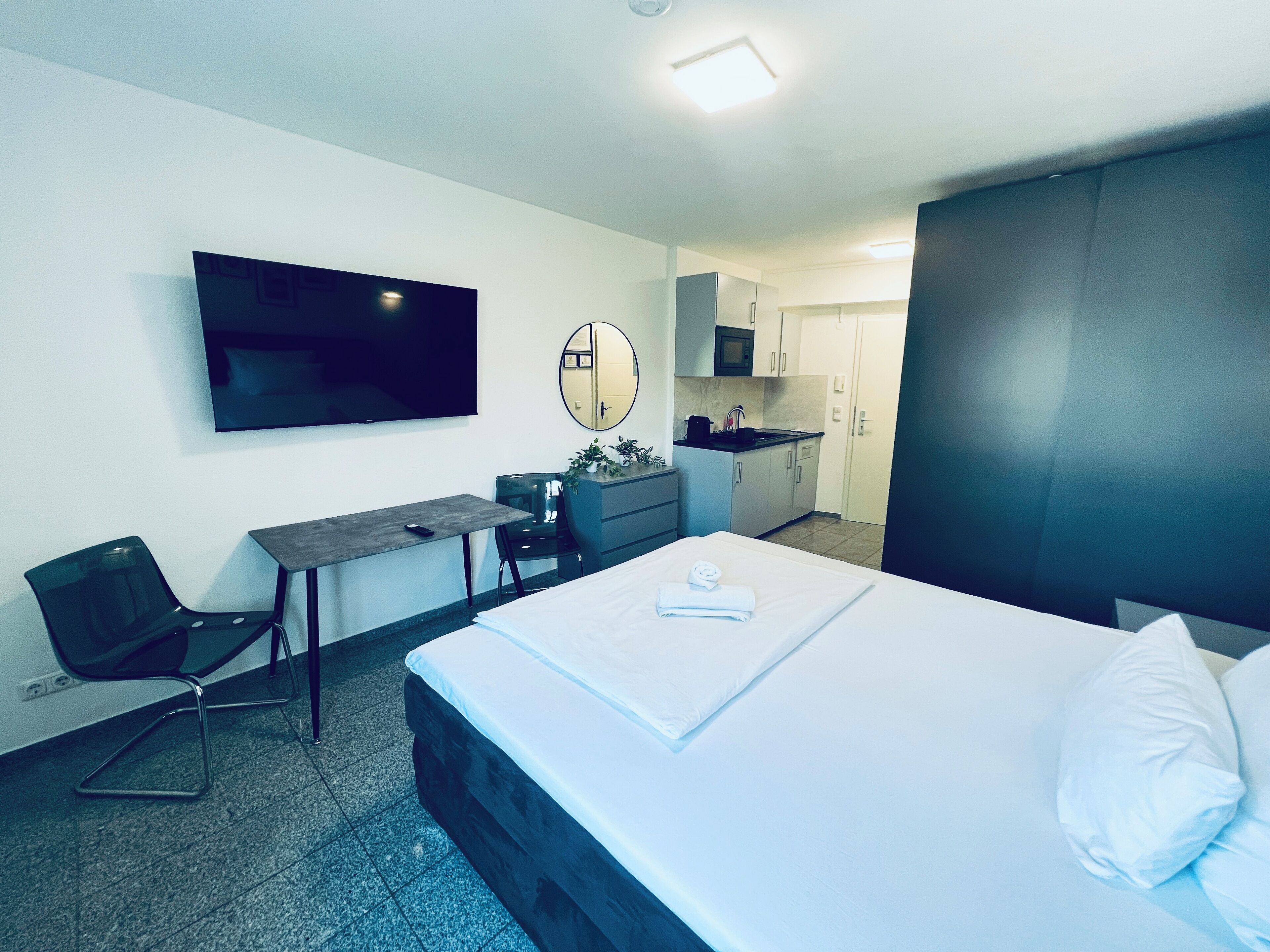 A hotel room with a bed, a desk, a chair, a TV, and a kitchen area.