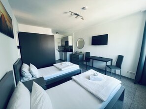 A modern hotel room with two beds, a desk, a chair, a TV, and a kitchenette.