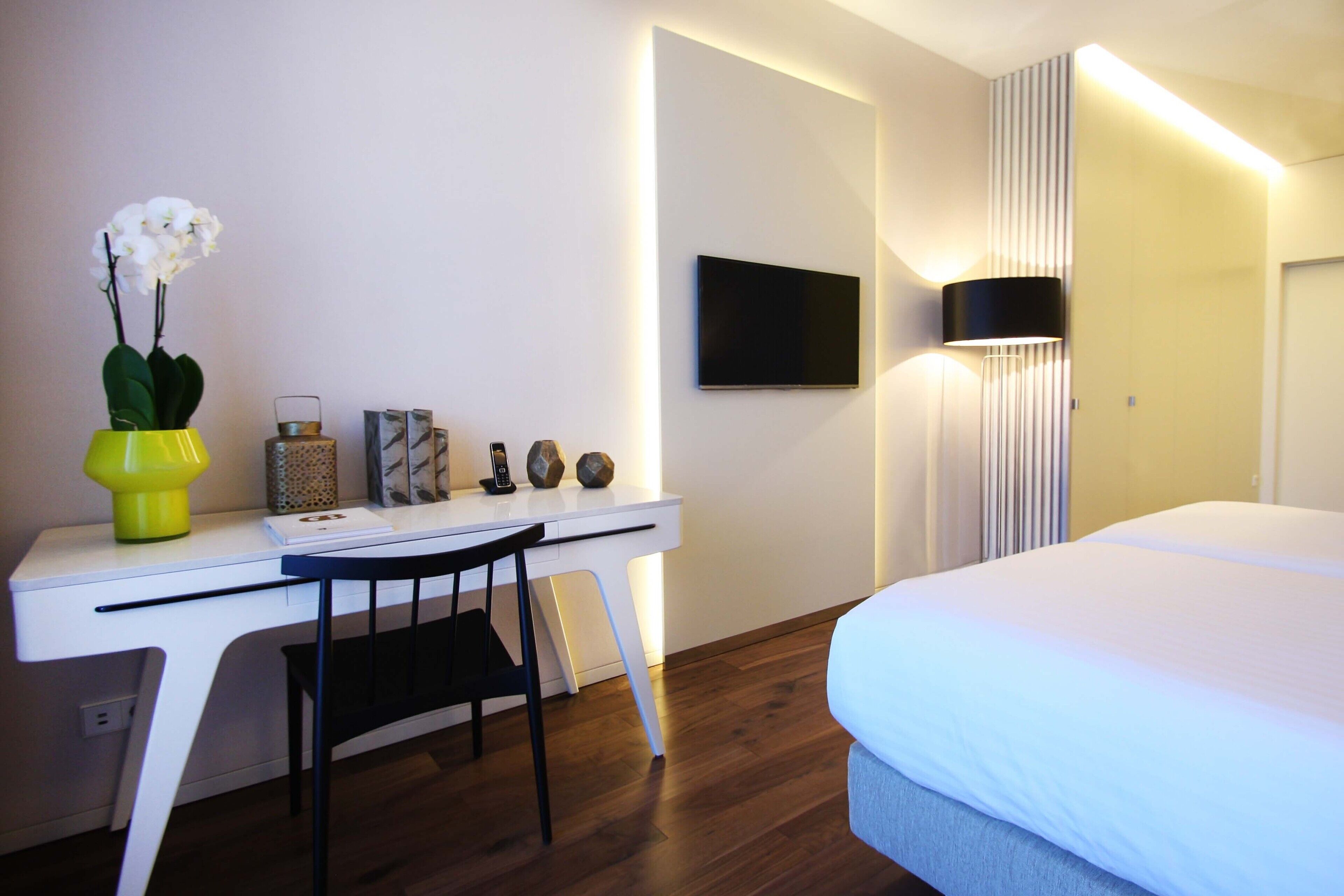 Una camera d'albergo con una scrivania bianca, una sedia, un letto, una TV a schermo piatto montata a parete e una lampada da comodino.