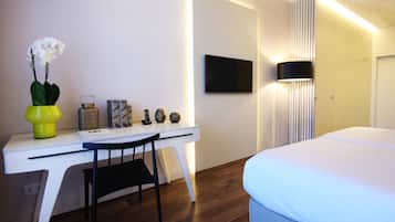 Habitación de hotel con un escritorio blanco, una silla, una cama, un televisor de pantalla plana montado en la pared y una lámpara de noche.