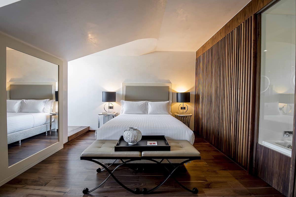 A modern hotel room with a large bed, bedside tables, a bedside lamp, a tray with a vase, and a wooden panel wall.