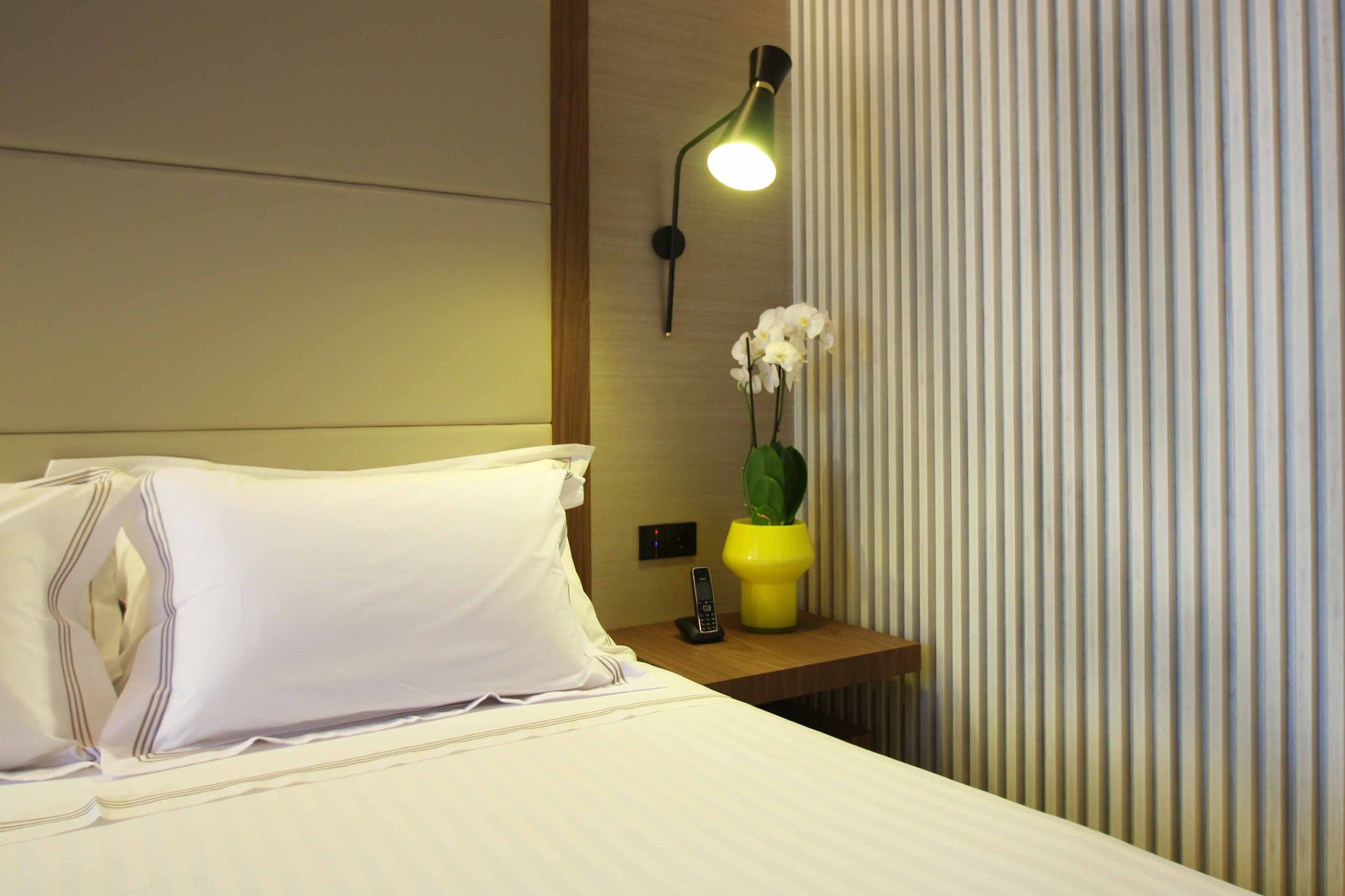 A neatly made bed with white linens, a bedside table with a phone and a vase with flowers, and a wall-mounted lamp.