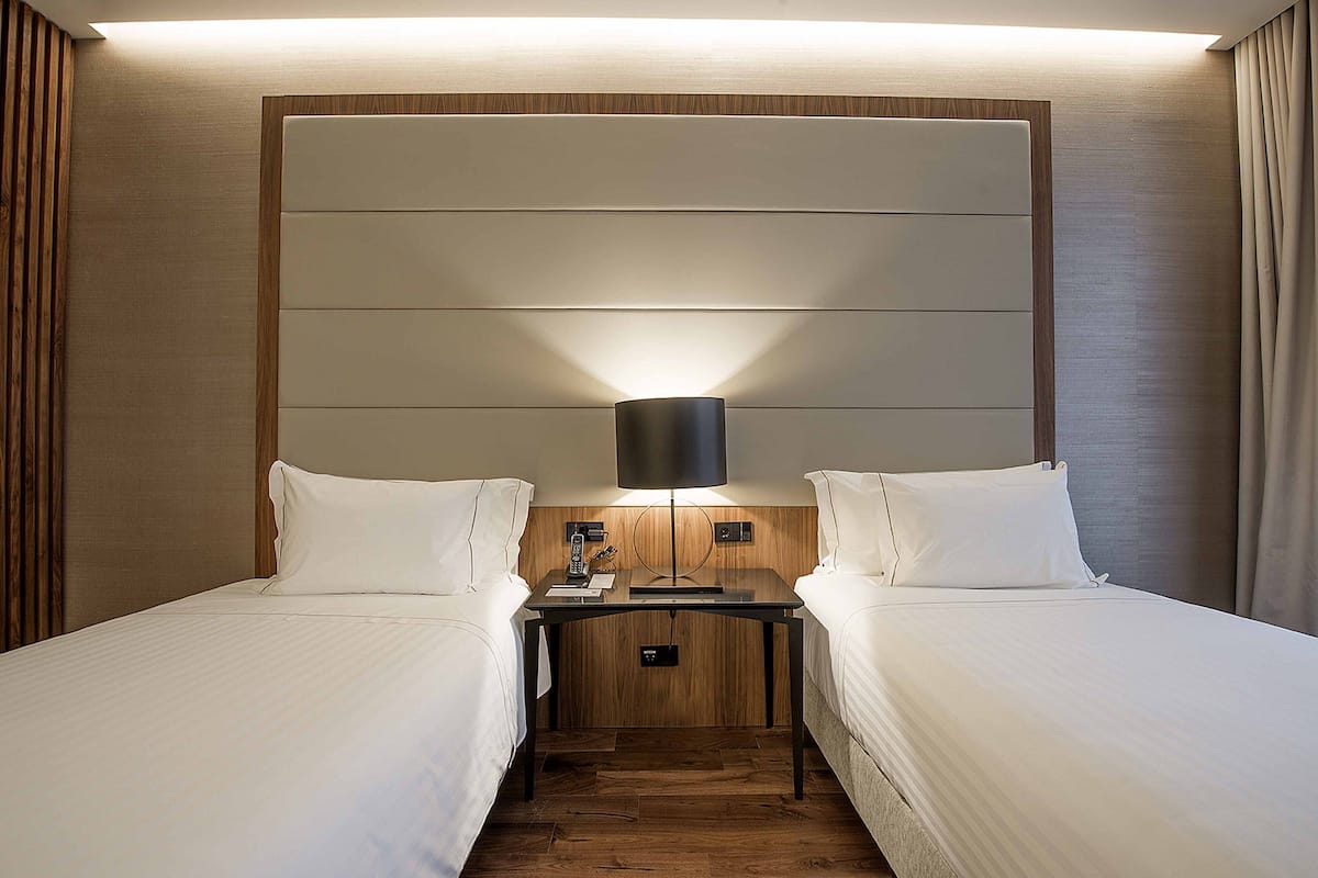 A hotel room with two beds, a nightstand, and a wall-mounted lamp.