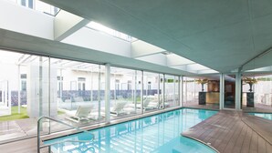 Una piscina interior moderna con paredes de vidrio transparente, una terraza de madera y sillones de descanso.