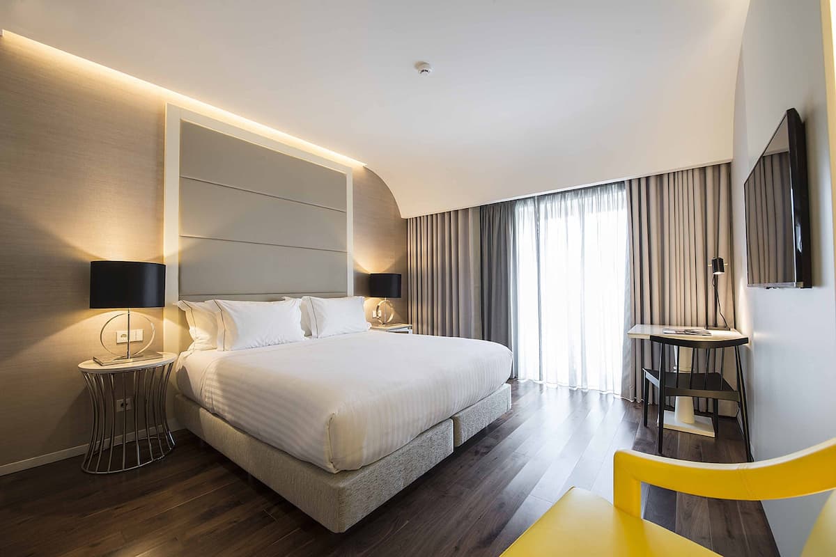 A modern hotel room with a large bed, bedside tables with lamps, a desk with a chair, and a large mirror.