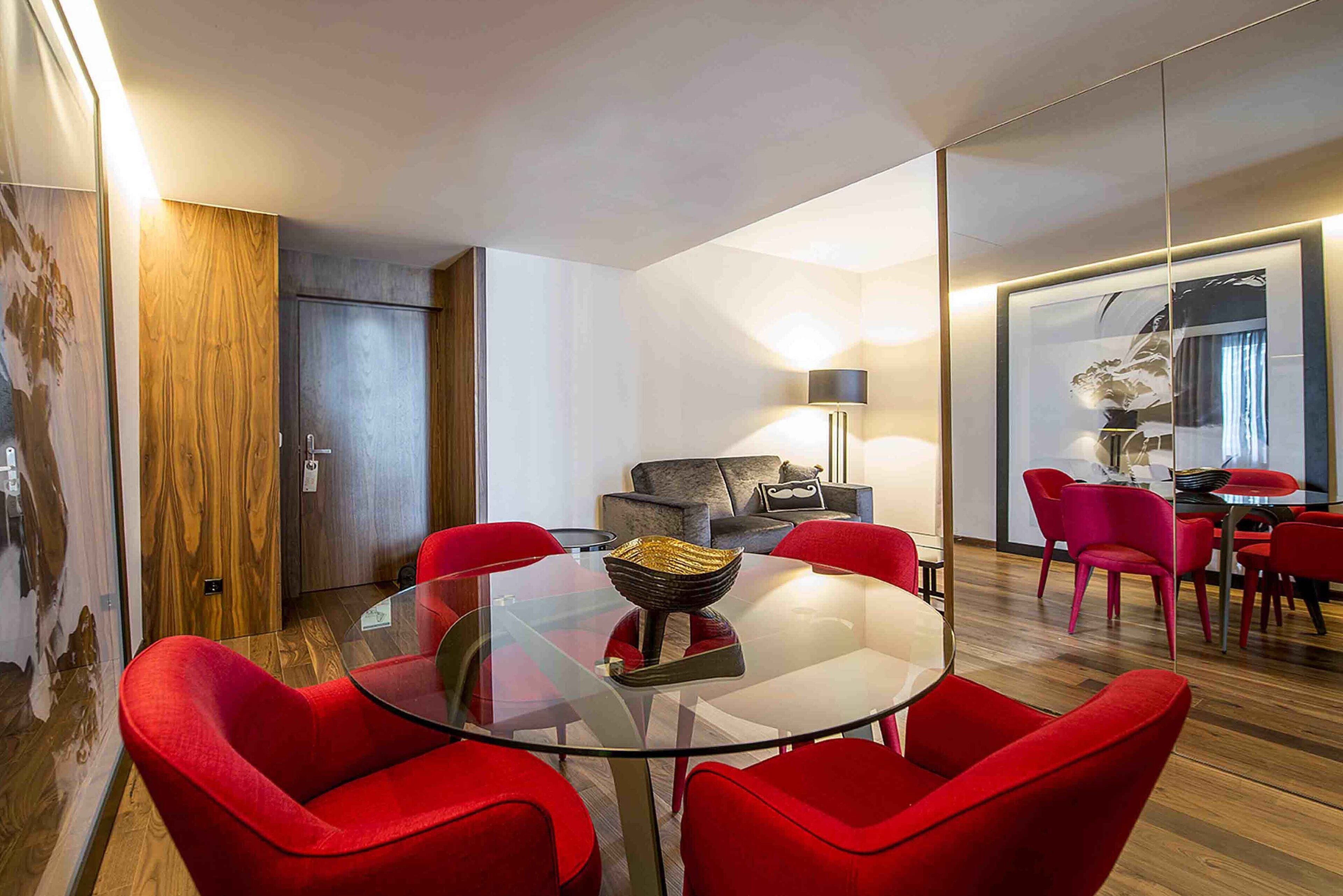 A modern living room with a glass table surrounded by red chairs, a wooden door, and a sofa with a lamp.