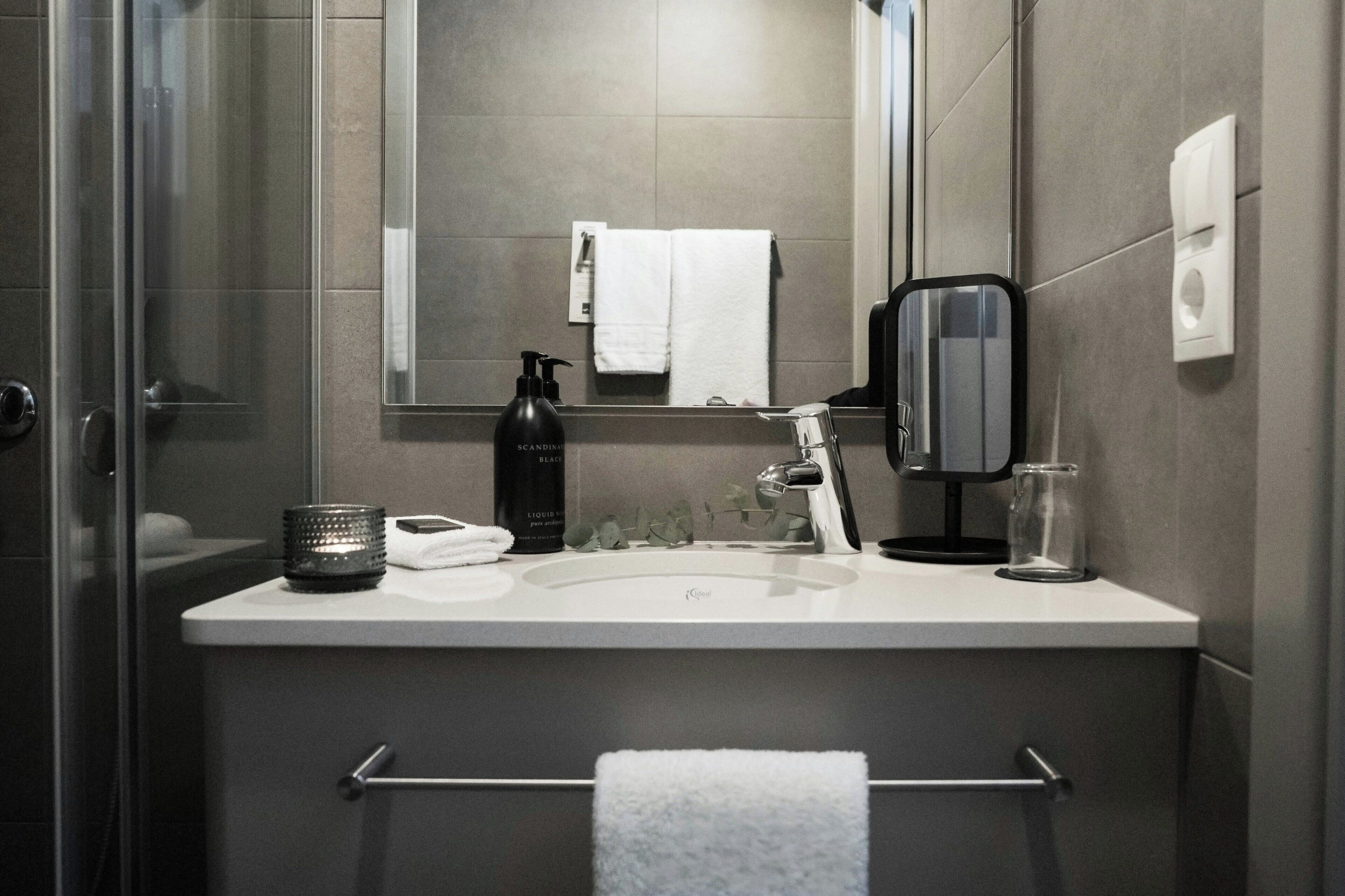 Un baño moderno con cabina de ducha de vidrio, un lavabo blanco, un espejo, un perchero para toallas, un dispensador de jabón y un vaso de agua.