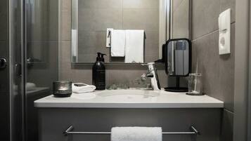 Un baño moderno con cabina de ducha de vidrio, un lavabo blanco, un espejo, un perchero para toallas, un dispensador de jabón y un vaso de agua.