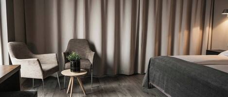 Una habitación de hotel moderna con una cama, dos sillones, una mesita redonda con una planta y un gran ventanal con cortinas.
