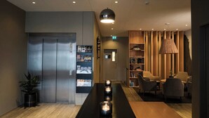 A modern lobby with a reception desk, a long table with candles, a bookshelf, and a seating area with chairs.