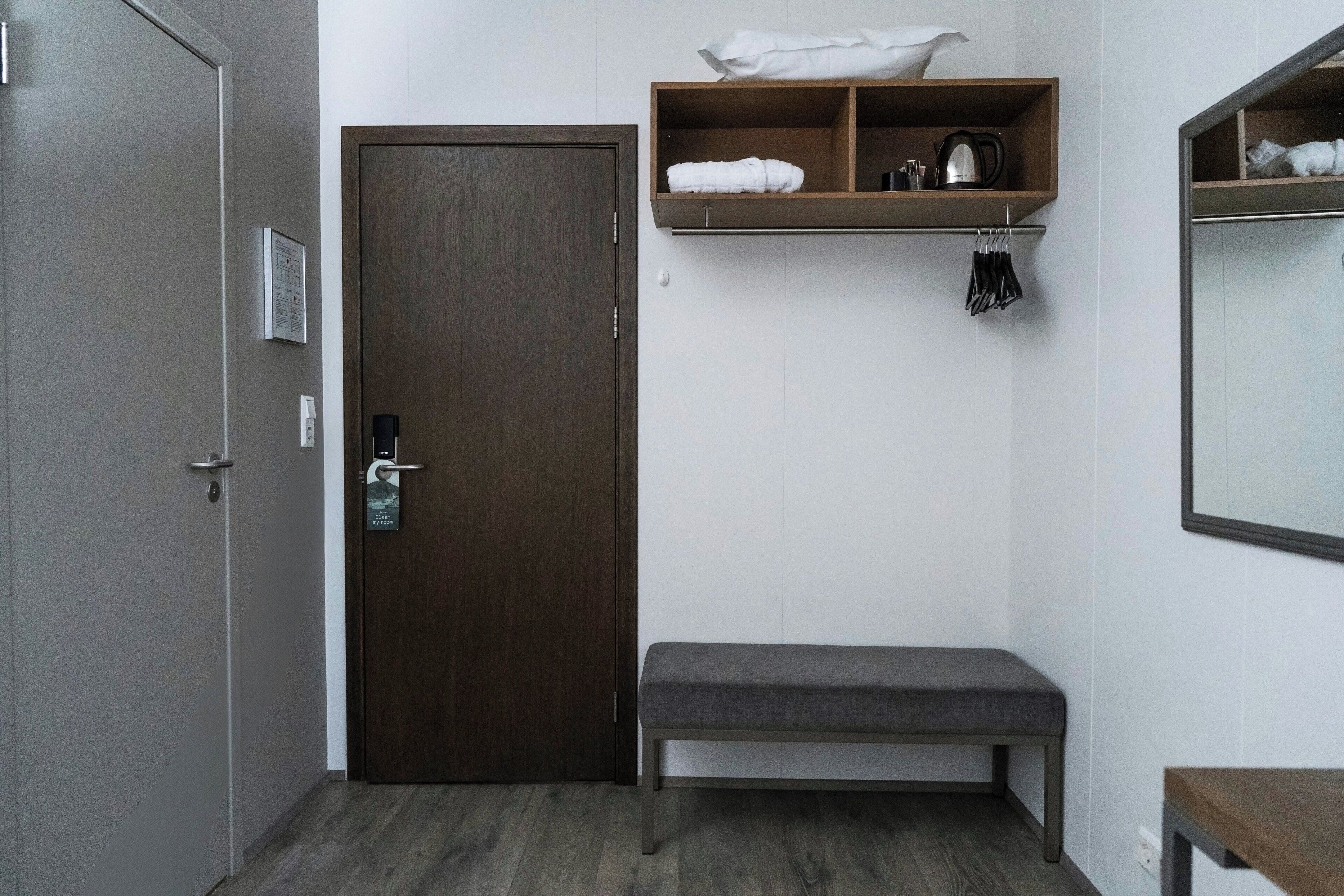 A hotel room with a wooden door, a bench, a shelf with towels and toiletries, and a mirror.