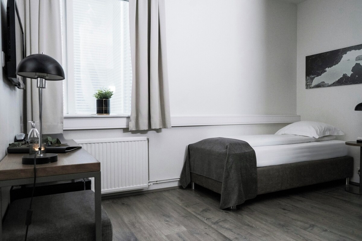 A minimalist bedroom with a single bed, a desk with a lamp, a radiator, and a window with curtains.