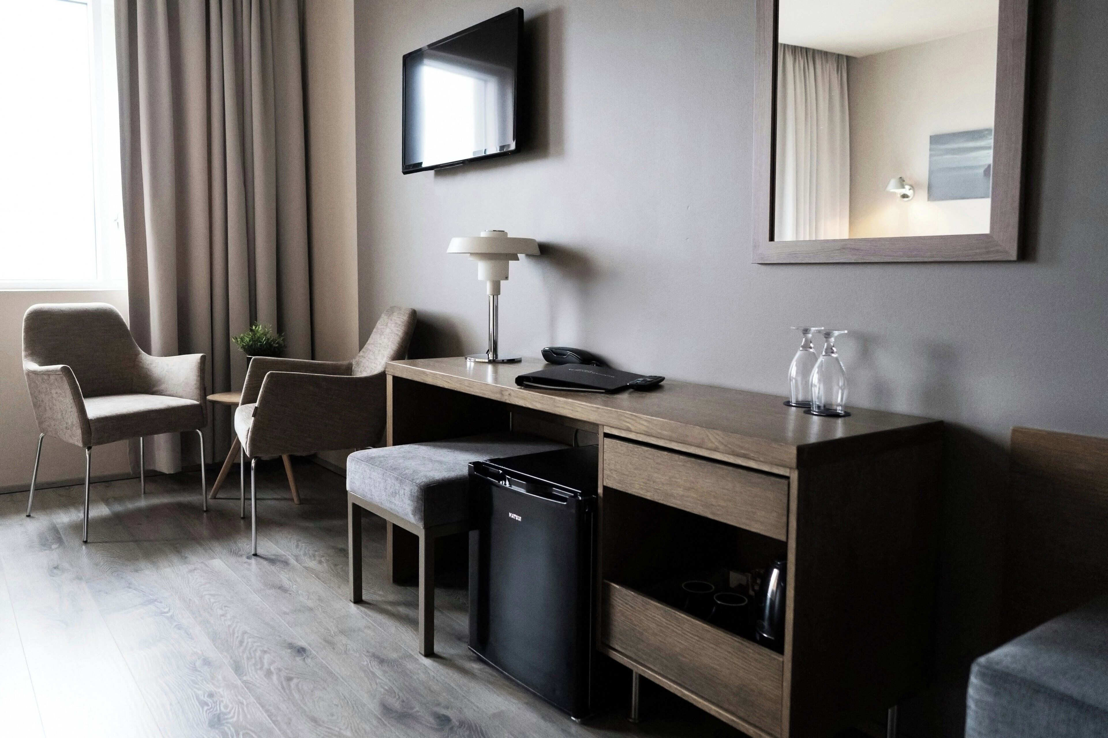 A modern hotel room with a wooden desk, two chairs, a small black refrigerator, a desk lamp, a flat-screen TV, and a mirror.