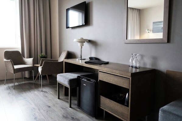 A modern hotel room with a wooden desk, two chairs, a small black refrigerator, a desk lamp, a flat-screen TV, and a mirror. - Hotel Vestmannaeyjar (Vestmannaeyjar)