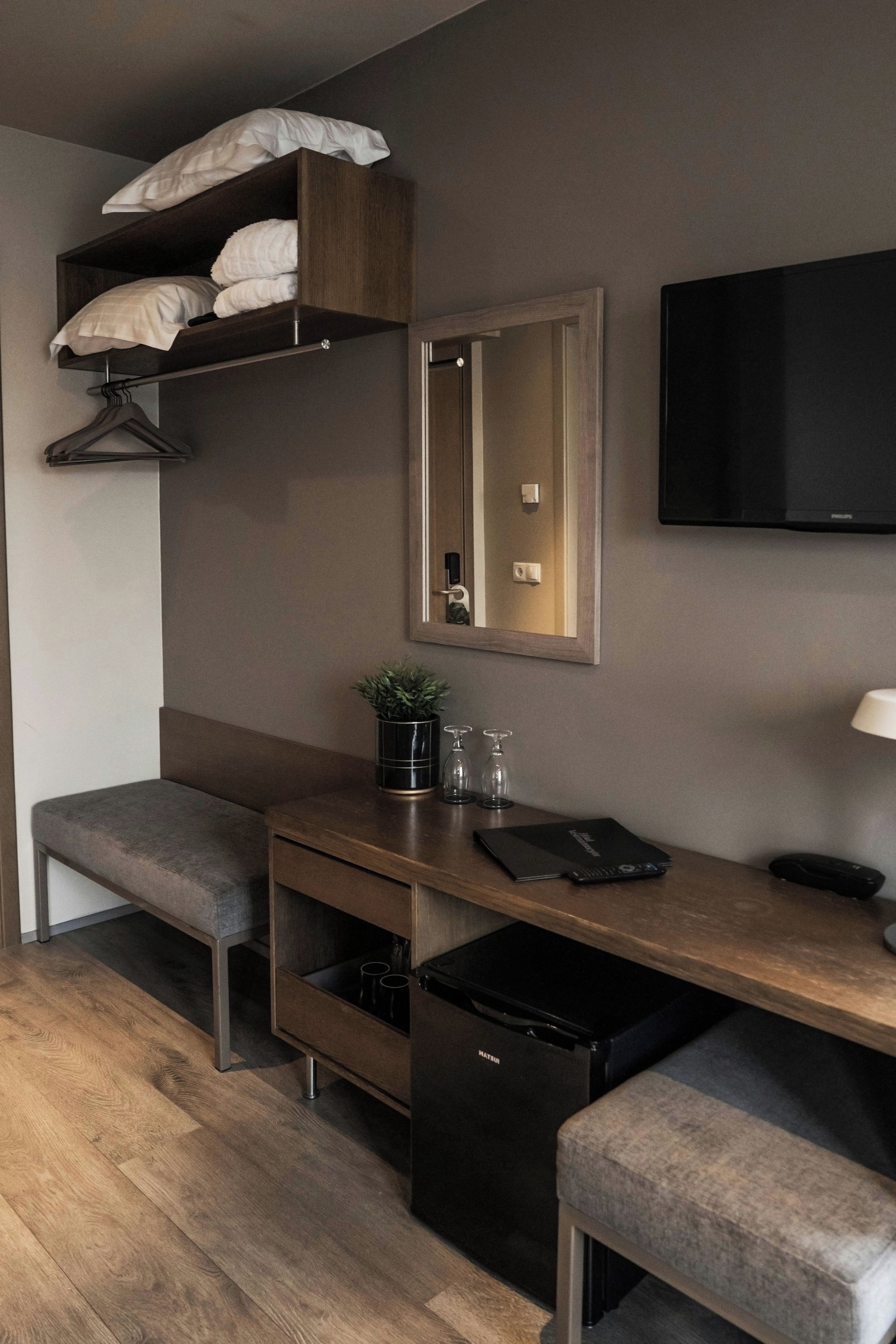 A compact hotel room with a wooden desk, a bench, a small refrigerator, a mirror, and a TV mounted on the wall.