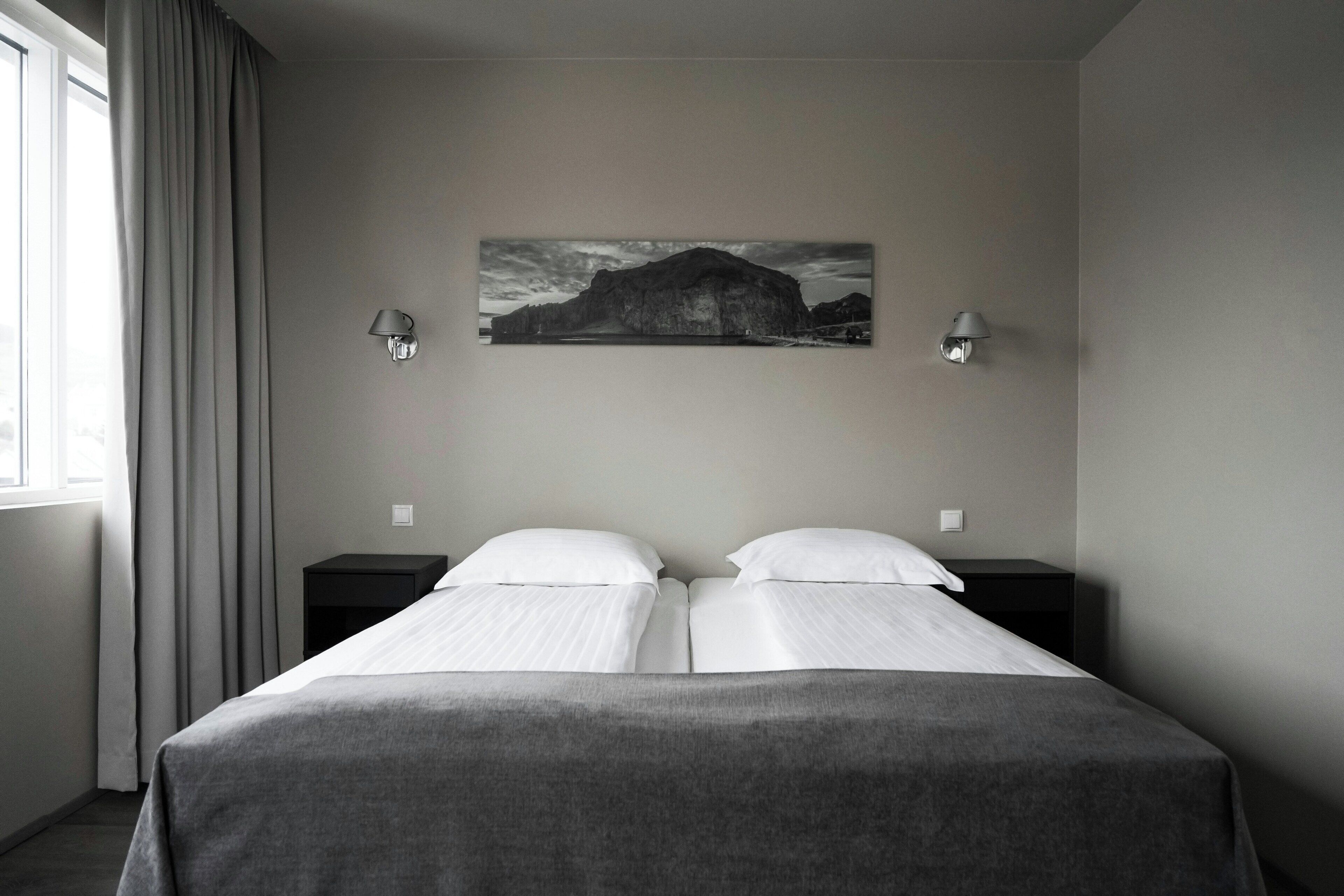 Habitación de hotel con una cama, dos mesitas de noche, una lámpara de pared y un cuadro de un paisaje montañoso.