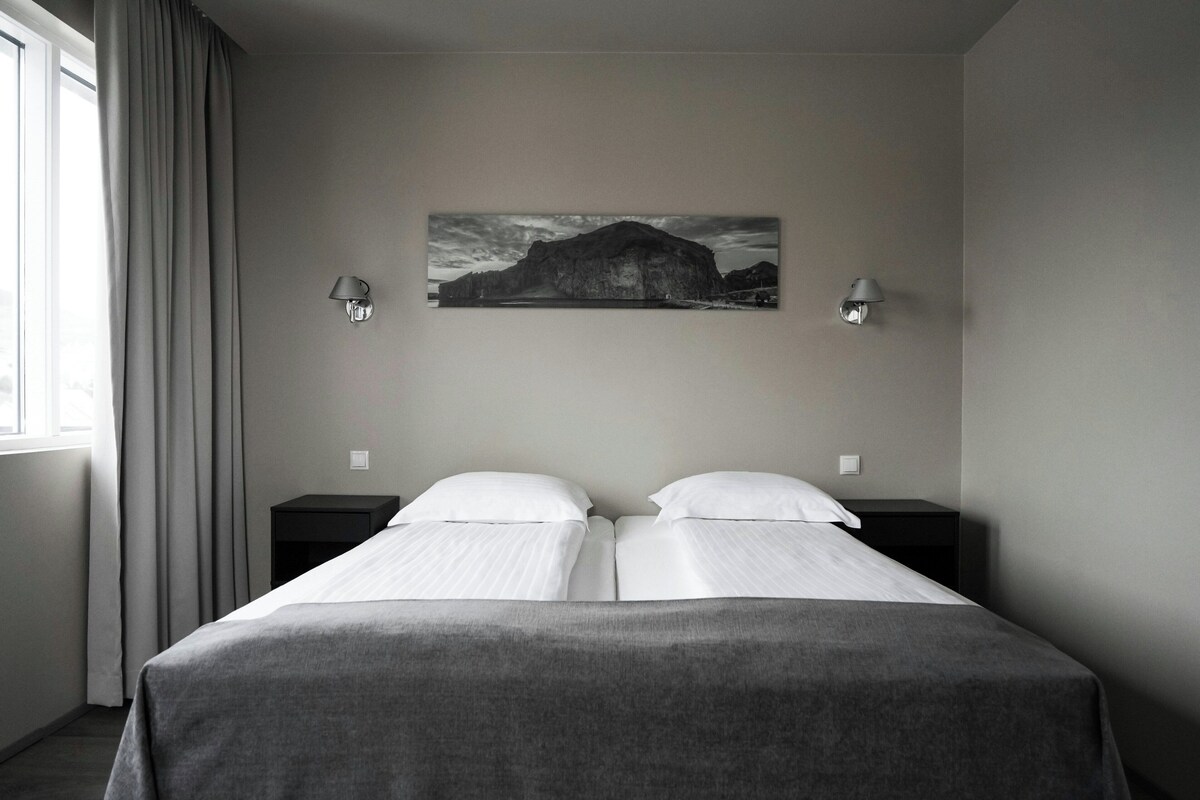 A hotel room with a bed, two bedside tables, a wall-mounted lamp, and a painting of a mountain landscape.