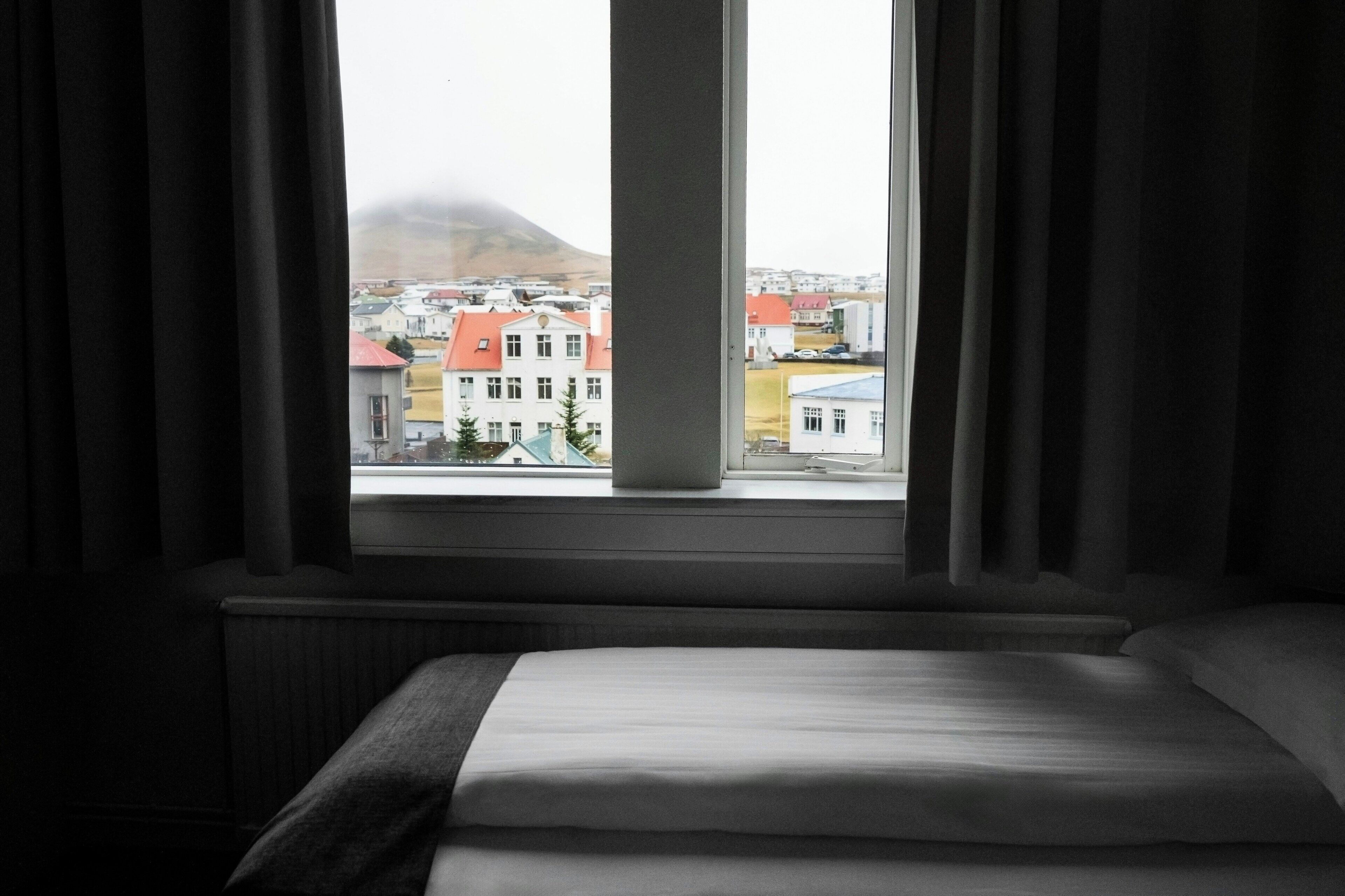 A hotel room with a bed, a window with curtains, and a view of buildings outside.