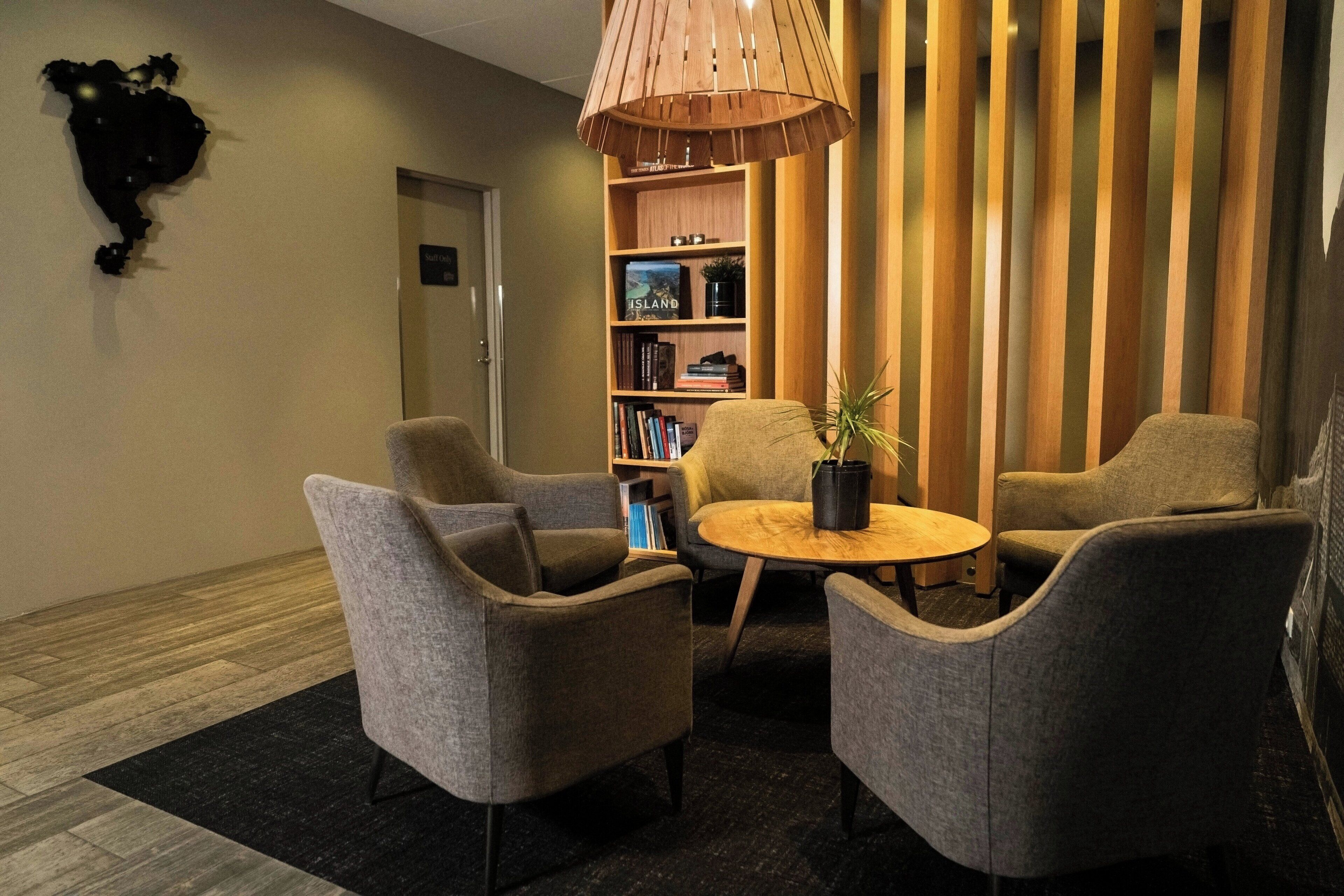 A modern lobby with grey armchairs, a round wooden table, a bookshelf, and a wooden slat partition.