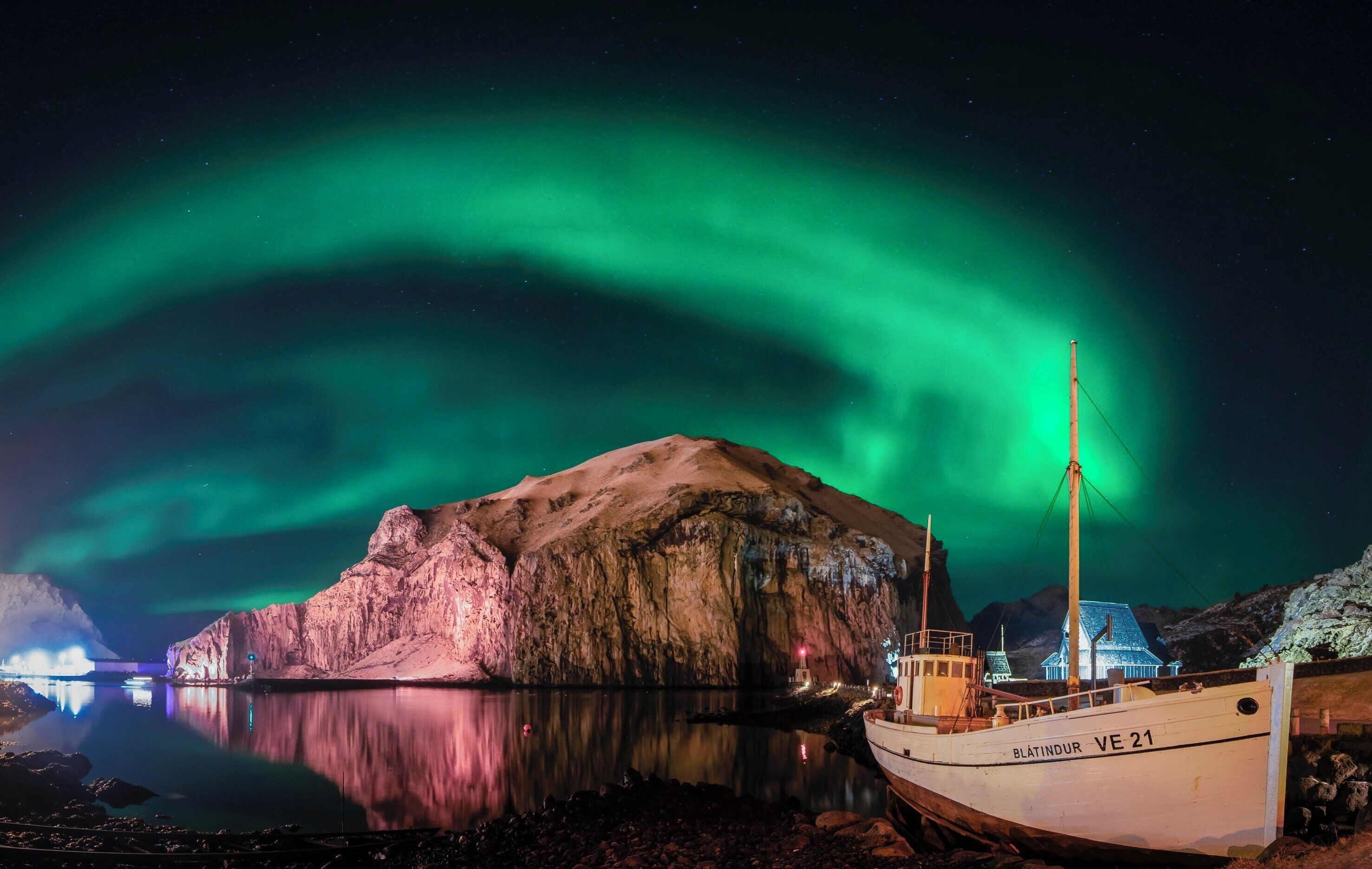 Un bote llamado "VE 21" atracado en una costa rocosa con un edificio grande e iluminado al fondo y la aurora boreal en el cielo.
